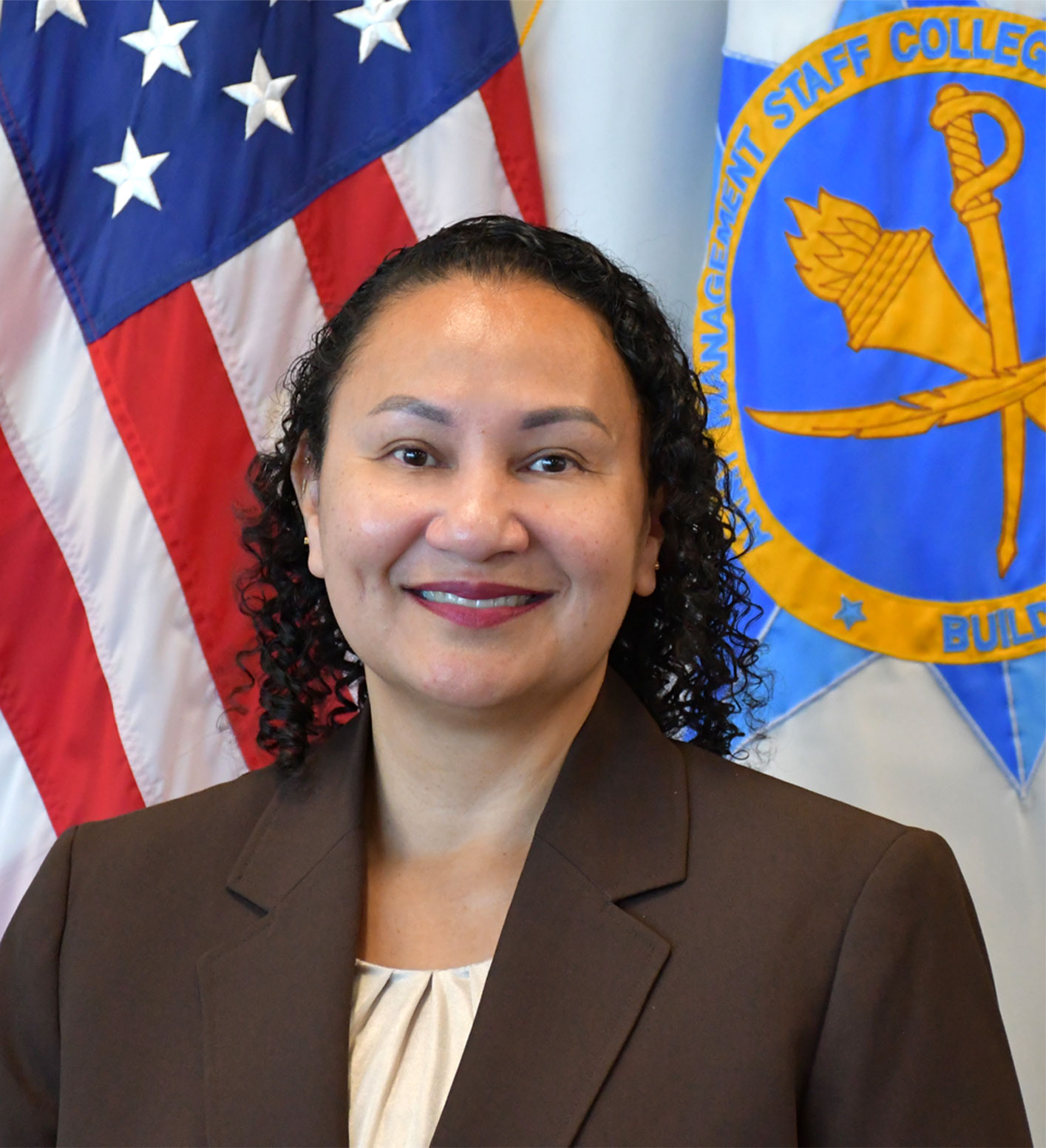 A person wearing a brown blazer stands in front of two flags: the United States flag and the Command and General Staff College flag featuring a torch and sword emblem.