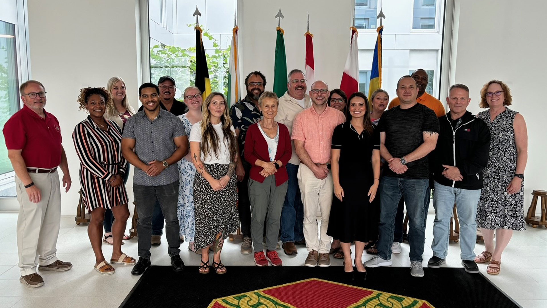A group of approximately twenty people—diverse in age, gender, and ethnicity—stand together indoors in front of several national flags, smiling for a group photo. A red and green emblem is visible on the floor.