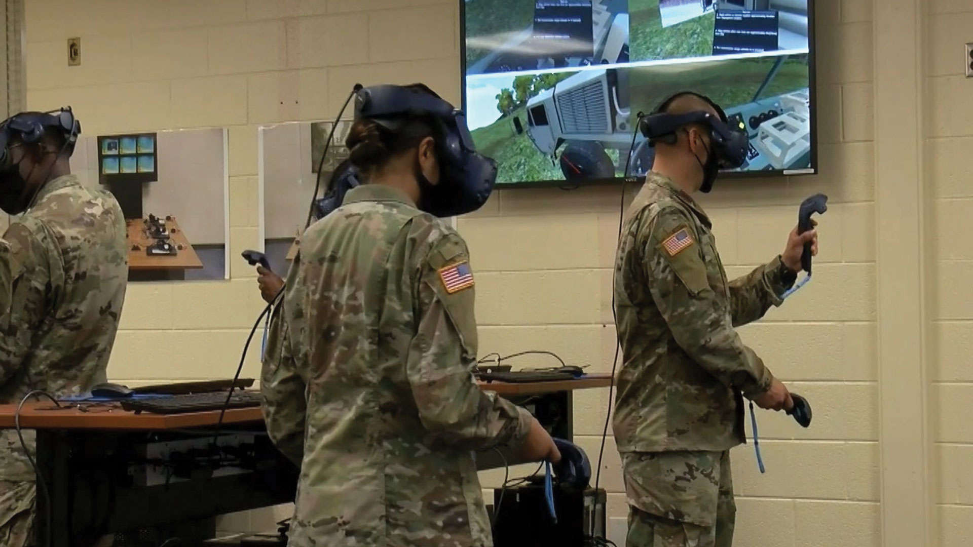 Soldiers in uniform using virtual reality headsets and hand controllers during a training exercise in a classroom setting, with training visuals displayed on a monitor.