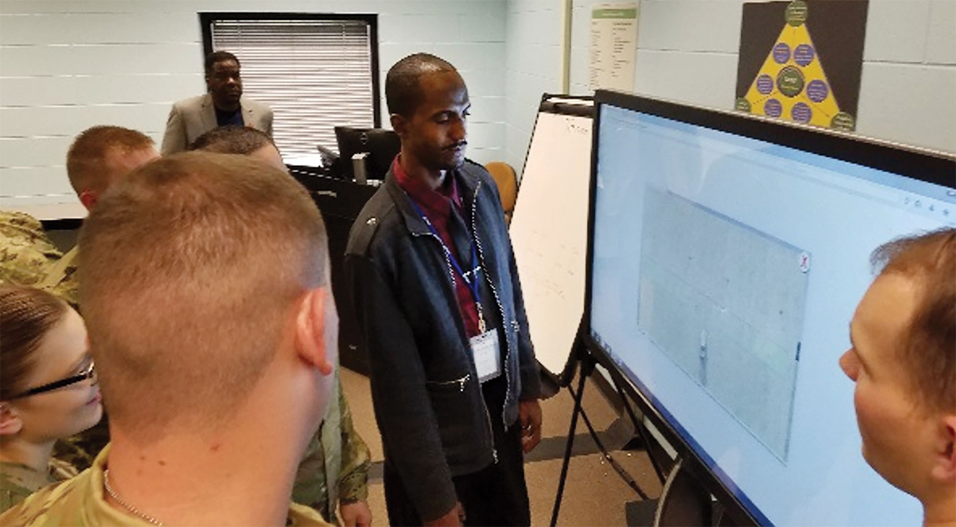 Army personnel gathered around a large interactive touchscreen display, viewing a 3D layout or map, while an instructor leads the session in a classroom.