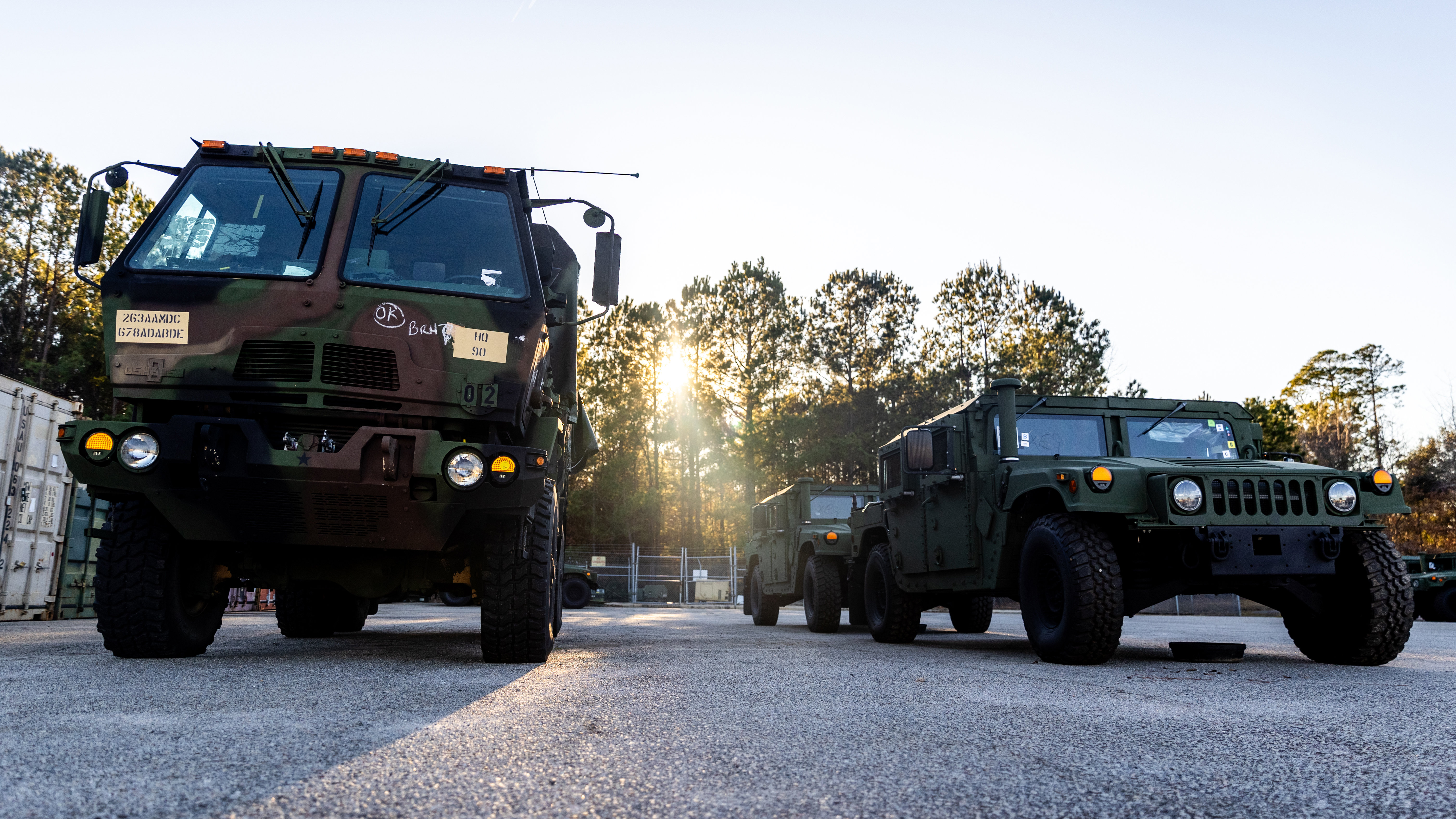 Two military vehicles parked on asphalt during golden hour, with sunlight filtering through trees in the background. On the left is a large tactical truck with camouflage paint and multiple antennas, while on the right is a smaller HMMWV (Humvee) utility vehicle. Both vehicles have their headlights on and are positioned in what appears to be a military facility or training area surrounded by pine trees.