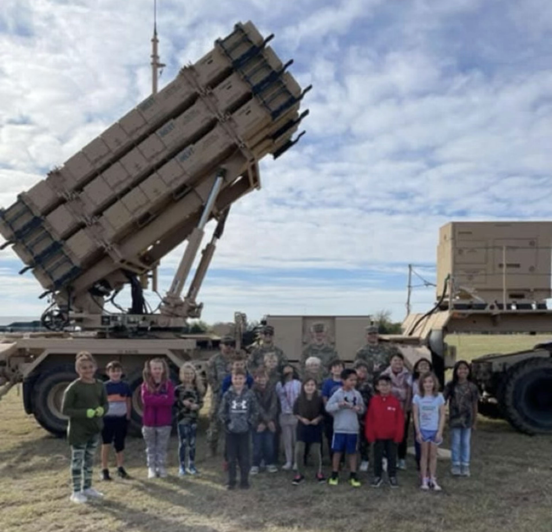 Group of children and soldiers standing in front of a large, elevated military missile launcher on a clear day.