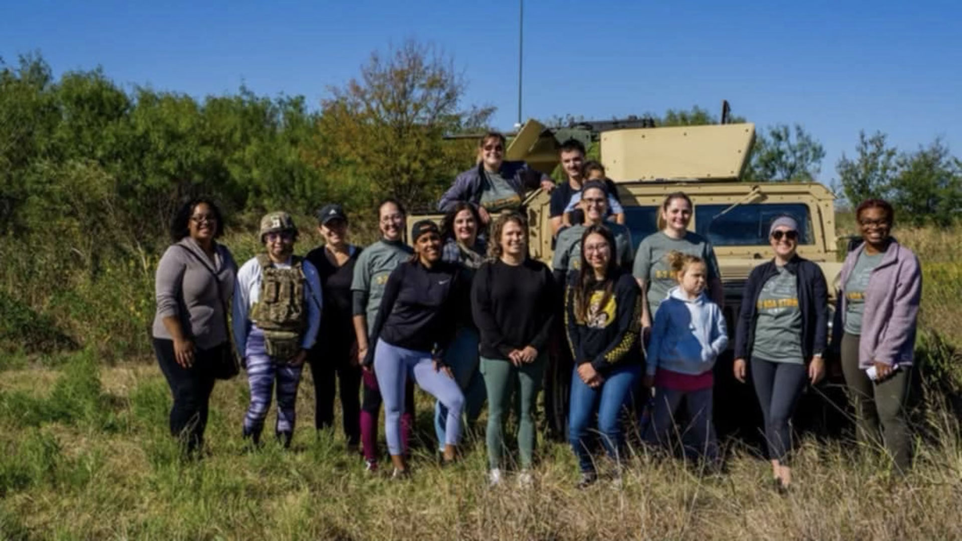 Group of women and military personnel posing in front of an armored military vehicle in a grassy field.