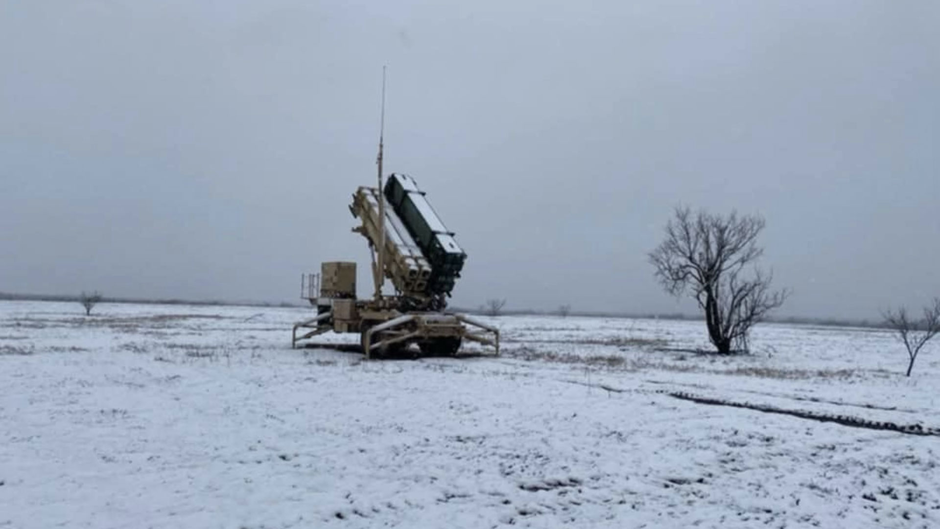 Military missile launcher stationed in a snow-covered field under overcast skies, with sparse trees nearby.