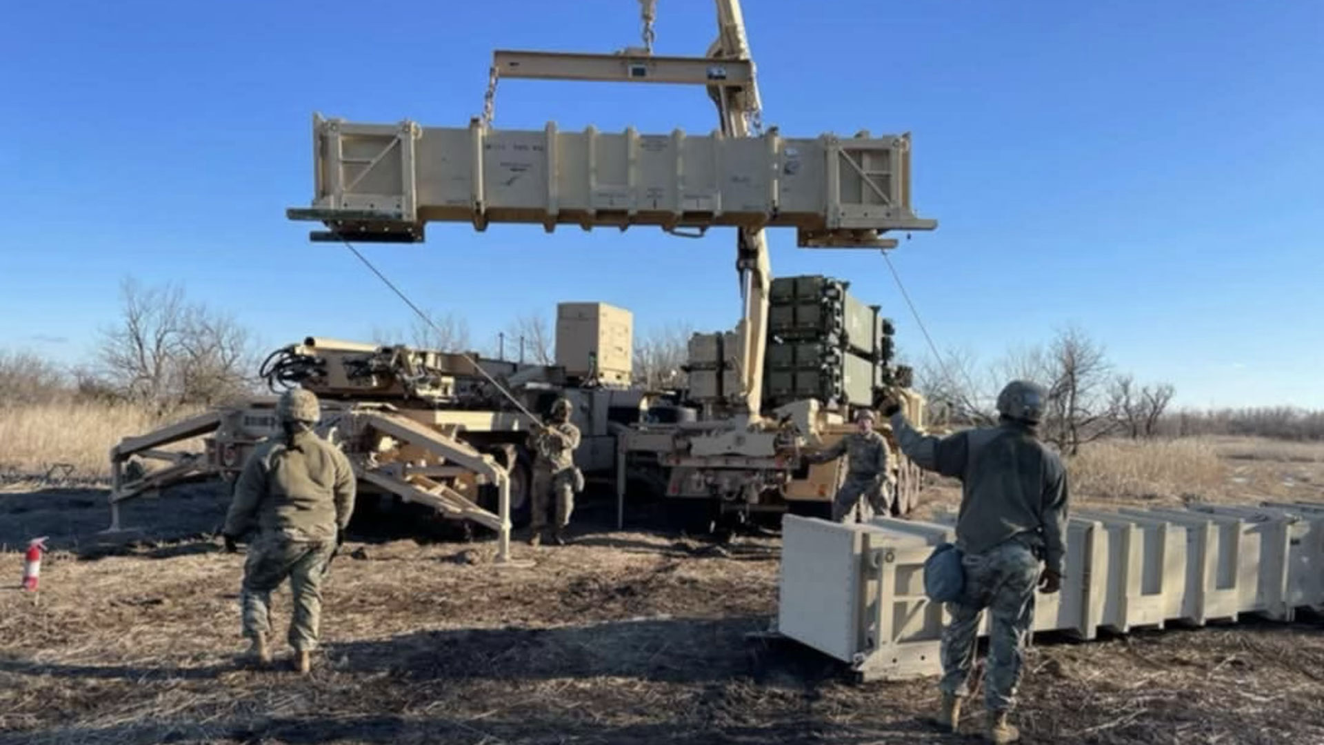 Soldiers working around a military launcher as a missile container is hoisted above by a crane in a dry, grassy field.
