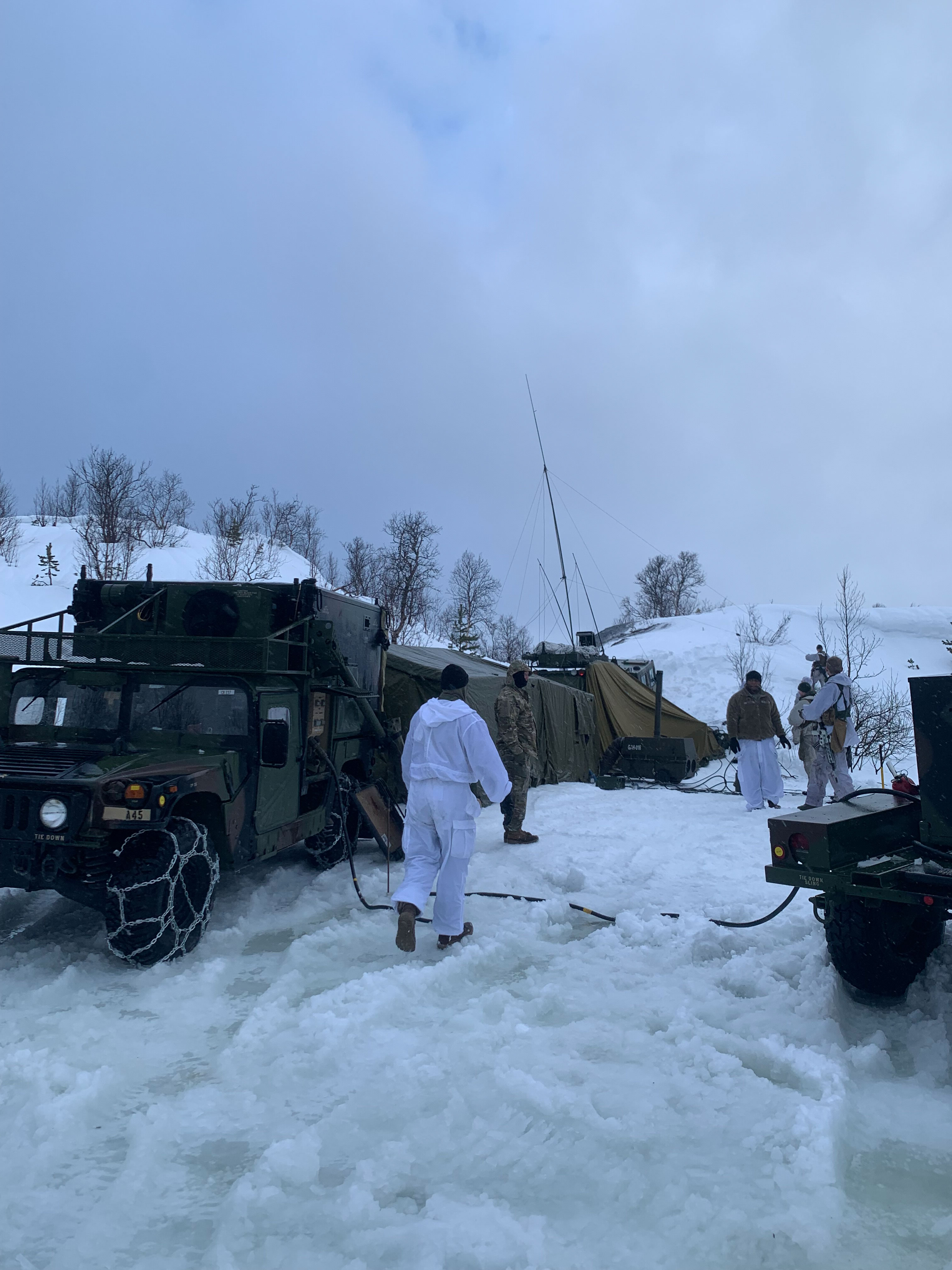 Soldiers in winter camouflage and cold-weather gear operate around a tactical command post with Humvees, antenna systems, and tents set up on a snow-covered landscape.