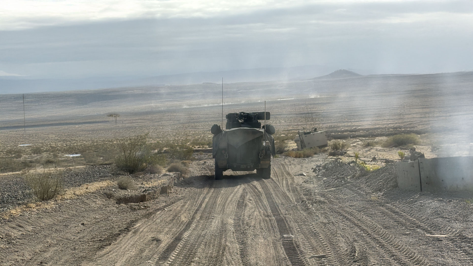 A military vehicle drives along a dusty desert trail, leaving deep tire tracks in the sand, with rugged terrain and distant mountains under a cloudy sky.