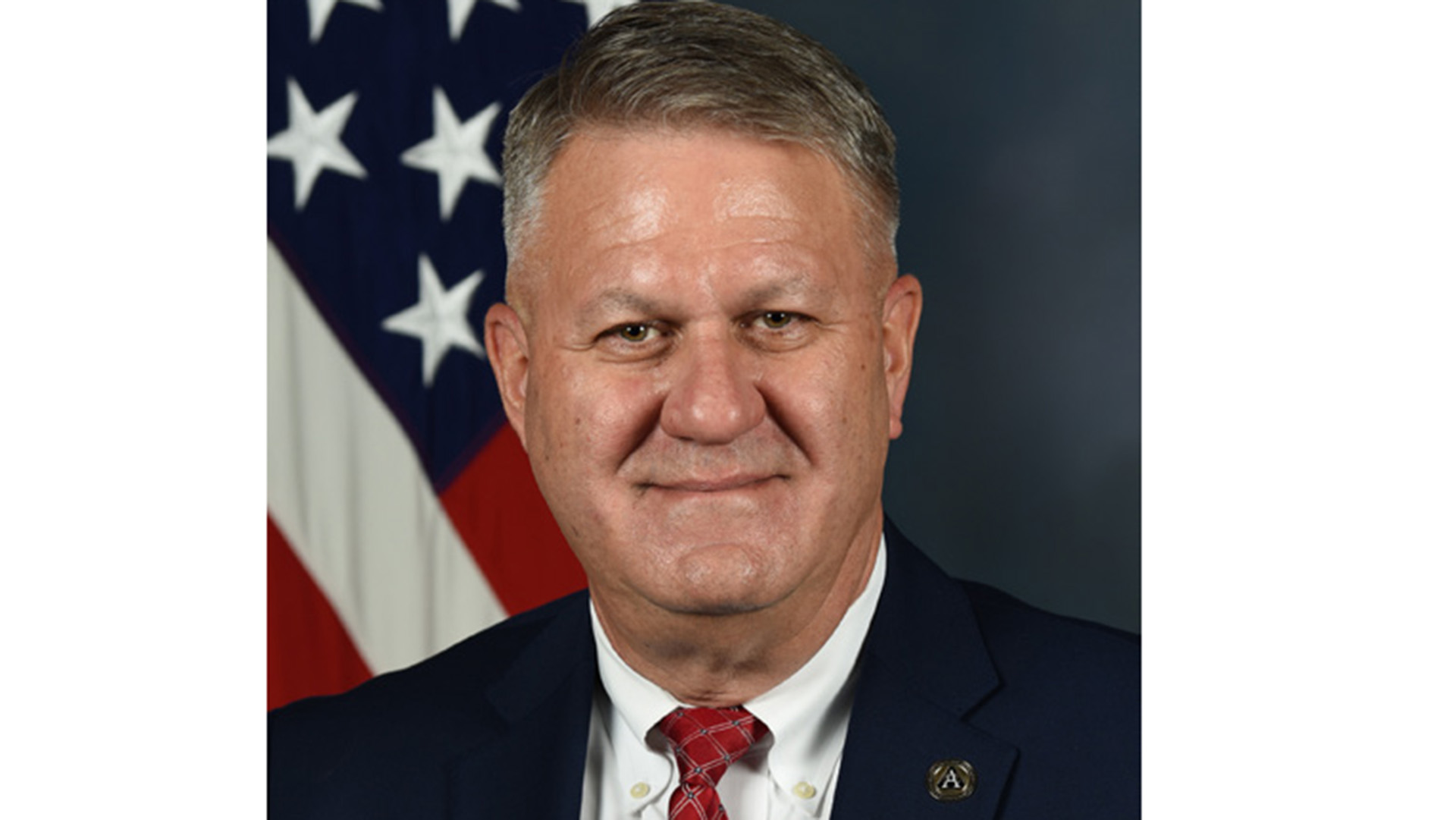 Headshot of a man in suit and red tie wearing a lapel pin in front of a U.S. flag