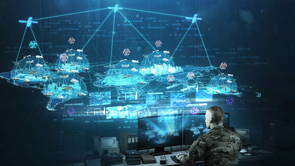 A military operator sits at a computer workstation viewing a futuristic blue holographic display showing satellite networks, data feeds, and interconnected systems with numerical data overlays. The visualization includes satellite icons and network connection lines spanning across what appears to be a global map.