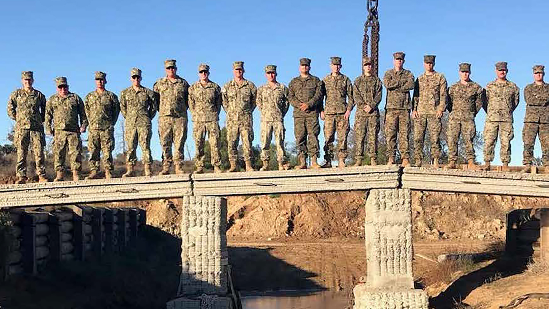 A formation of military personnel in desert camouflage uniforms standing at attention on a concrete bridge. The soldiers are lined up in a row across the bridge span, which crosses over a body of water that reflects both the bridge structure and the soldiers. The scene appears to be during a military ceremony or formal event.