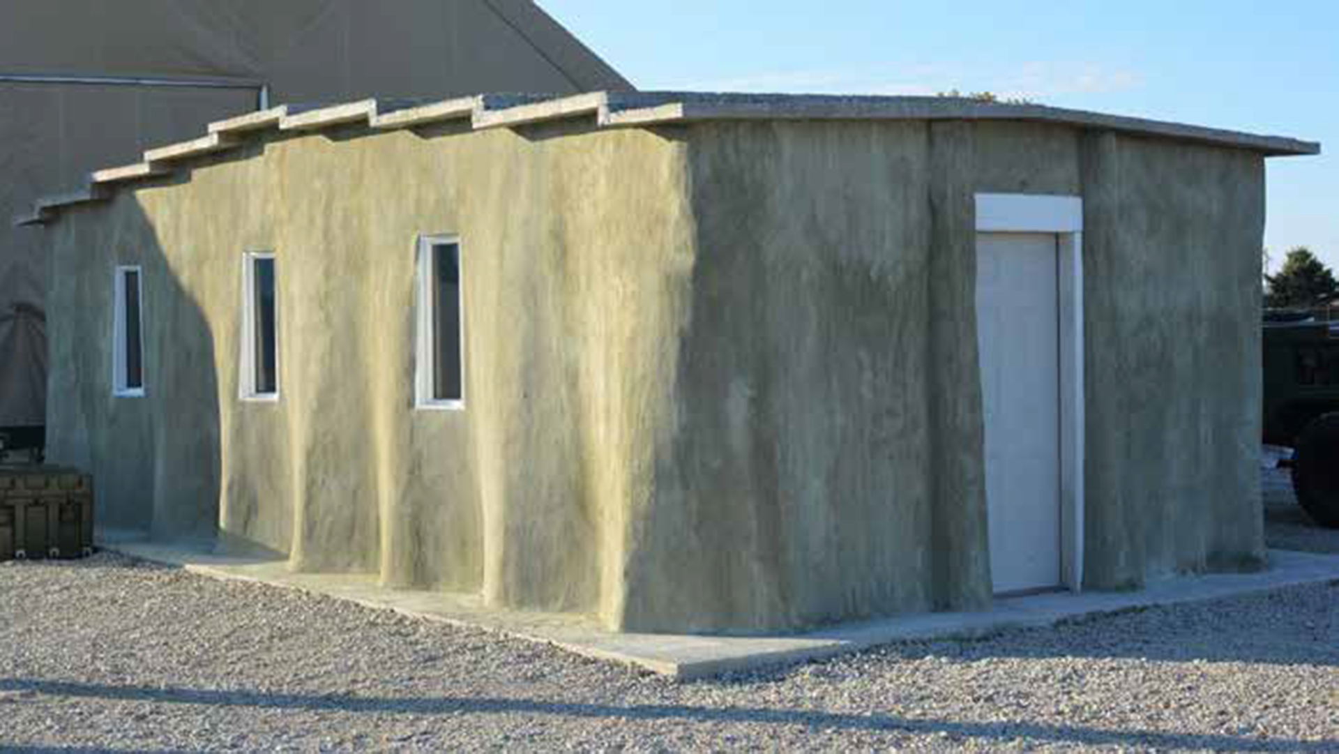 A single-story building with smooth concrete or stucco walls in gray and tan colors. The structure has several small rectangular windows and a white door, built on what appears to be a gravel foundation. The building has a simple, utilitarian design typical of military or temporary structures.