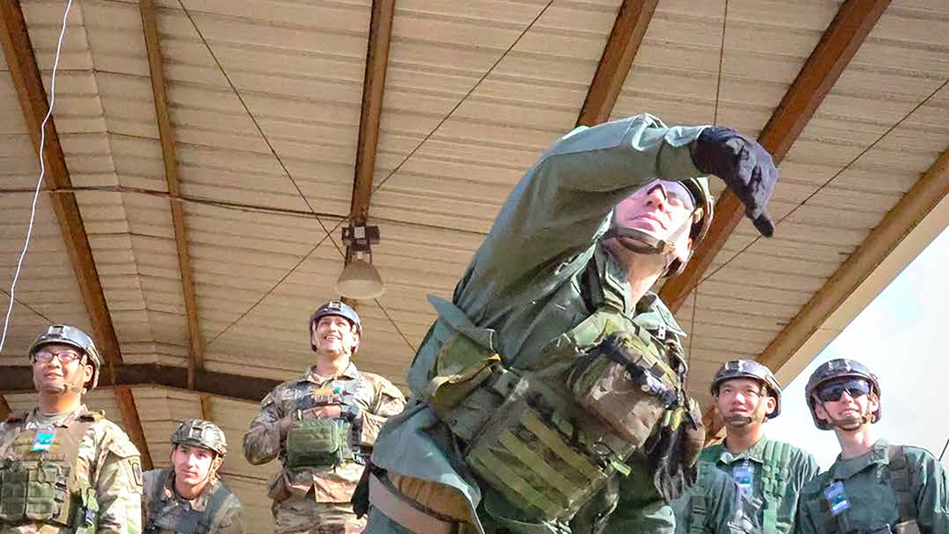 Military personnel in camouflage uniforms and helmets gathered around a soldier demonstrating proper lifting technique with a large military backpack in an indoor training facility