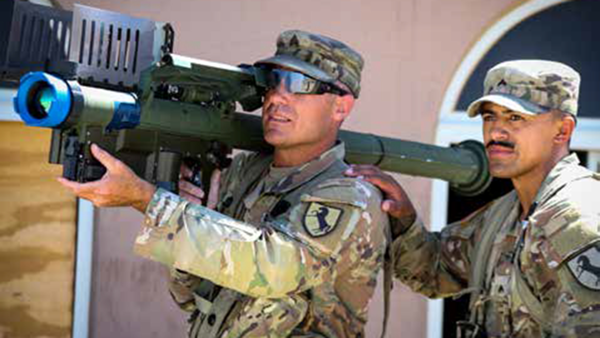 Two soldiers in desert camouflage uniforms examining a shoulder-fired weapon system, with one soldier holding the weapon while wearing sunglasses and a military cap