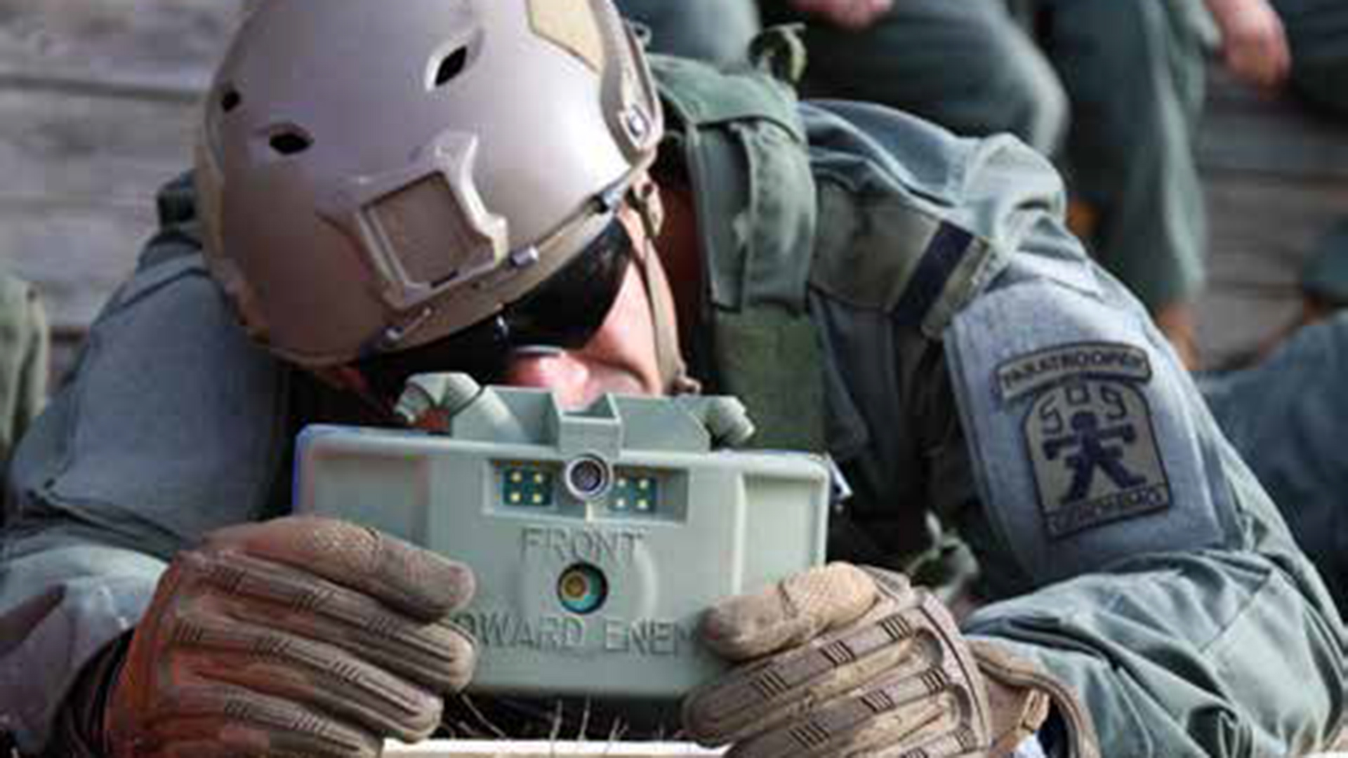 Soldier wearing protective helmet and gear using a handheld military device labeled 'FRONT TOWARD ENEMY' during field operations