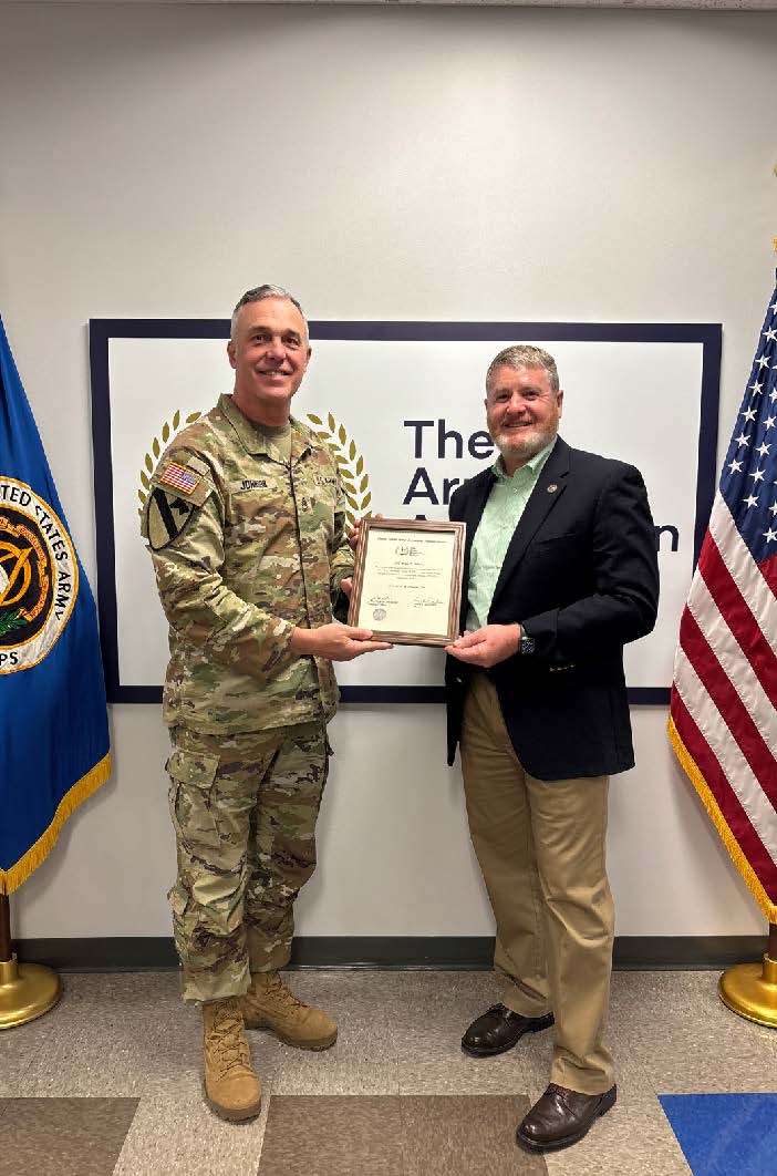 Two men stand side by side holding a framed certificate in front of a wall displaying The Army Acquisition School logo. On the left, Sgt. 1st Class Ryan Johnson wears Army combat uniform with a laurel wreath emblem visible on his chest. On the right, Craig Gardunia wears business casual attire with a light green shirt and dark blazer. American and Army flags frame both sides of the presentation.