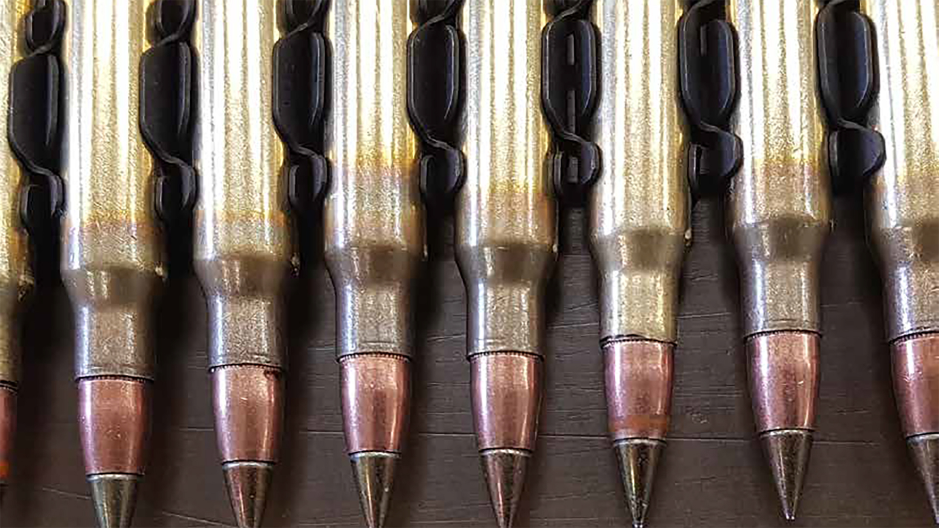 A row of rifle cartridges or ammunition rounds arranged in a line on a wooden surface. The cartridges have brass-colored casings with copper-colored bullet tips and black attachment points or clips connecting them together.