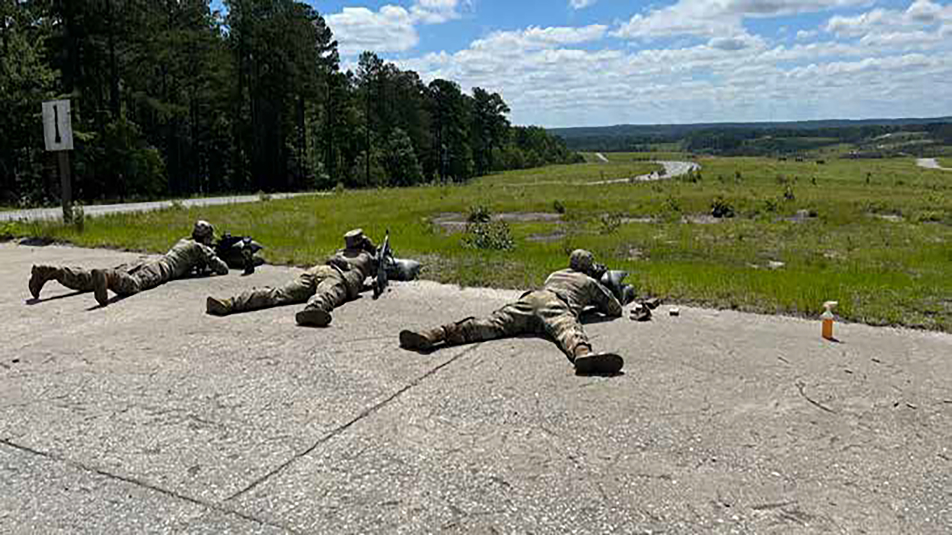 Military personnel in camouflage uniforms lying prone on a concrete surface during what appears to be a training exercise or drill. The soldiers are positioned in a firing line formation on what looks like a runway or large paved area, with trees and open fields visible in the background under a partly cloudy sky.