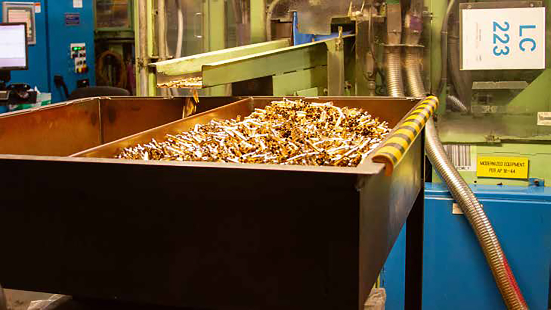 An industrial container or bin filled with thousands of spent brass cartridge casings in what appears to be a manufacturing or recycling facility. The brass casings are piled high in a large metal receptacle, with industrial equipment and machinery visible in the background.
