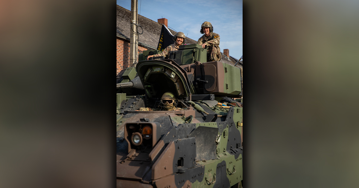 Three U.S. Army soldiers positioned on an armored vehicle, two standing on top while one is seated inside. They are wearing helmets and military gear, smiling and interacting with each other. The vehicle is painted in camouflage colors, and a flag with the words '1st Battalion' is visible in the background. Brick buildings and a clear sky provide the backdrop.