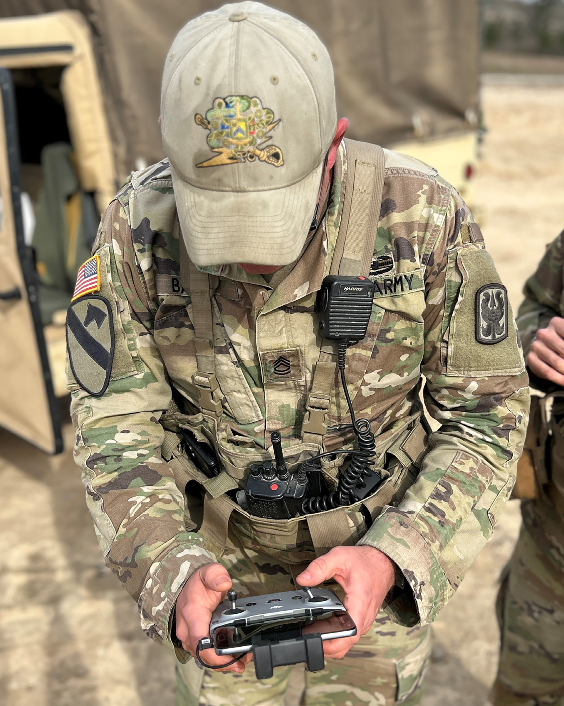 U.S. Army soldier in multicam uniform and tan baseball cap operating a drone controller, with radio equipment and patches visible on uniform, military vehicle in background