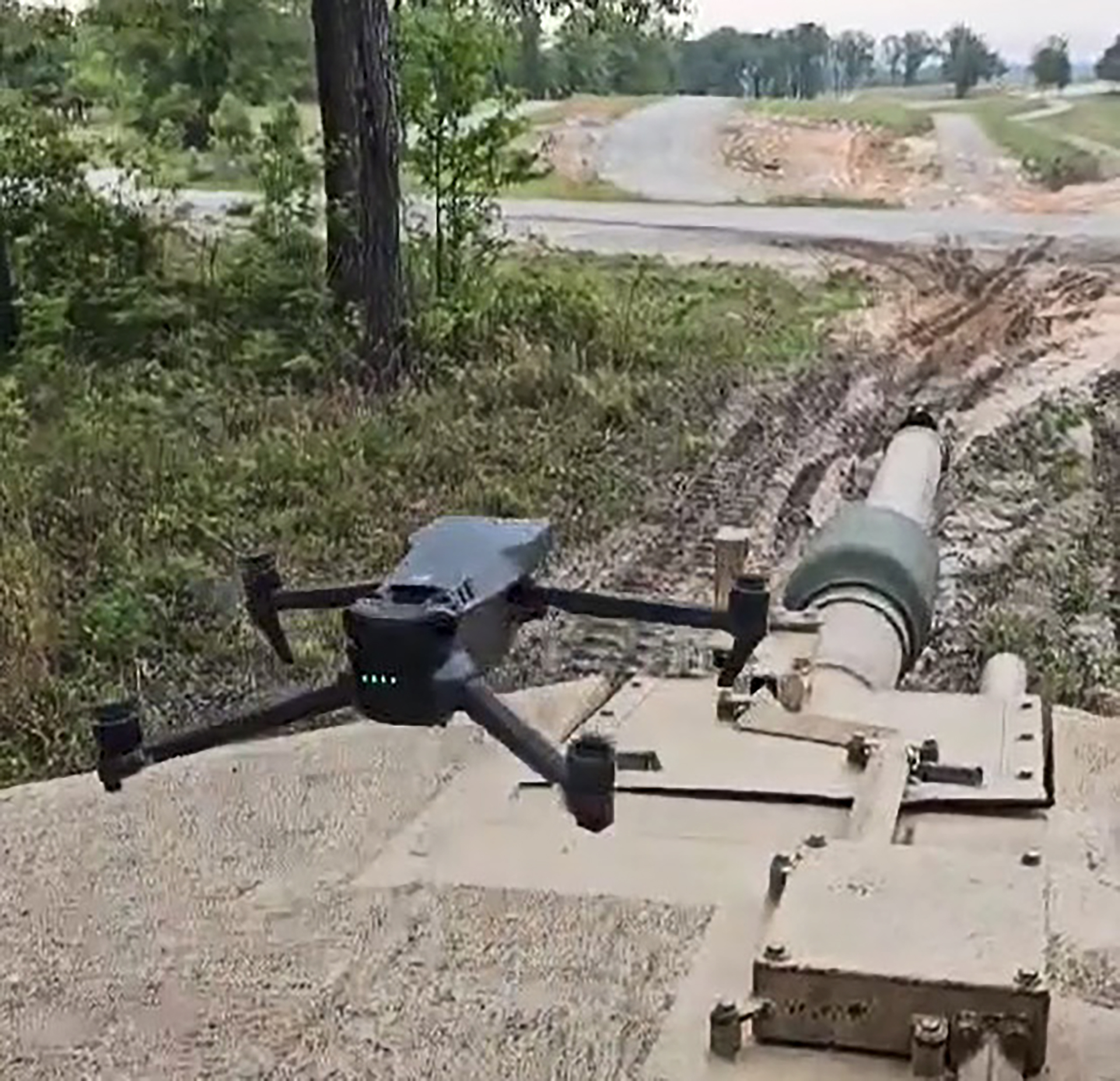 'Black quadcopter drone positioned on top of a military armored vehicle in an outdoor training area with dirt roads and vegetation visible in the background