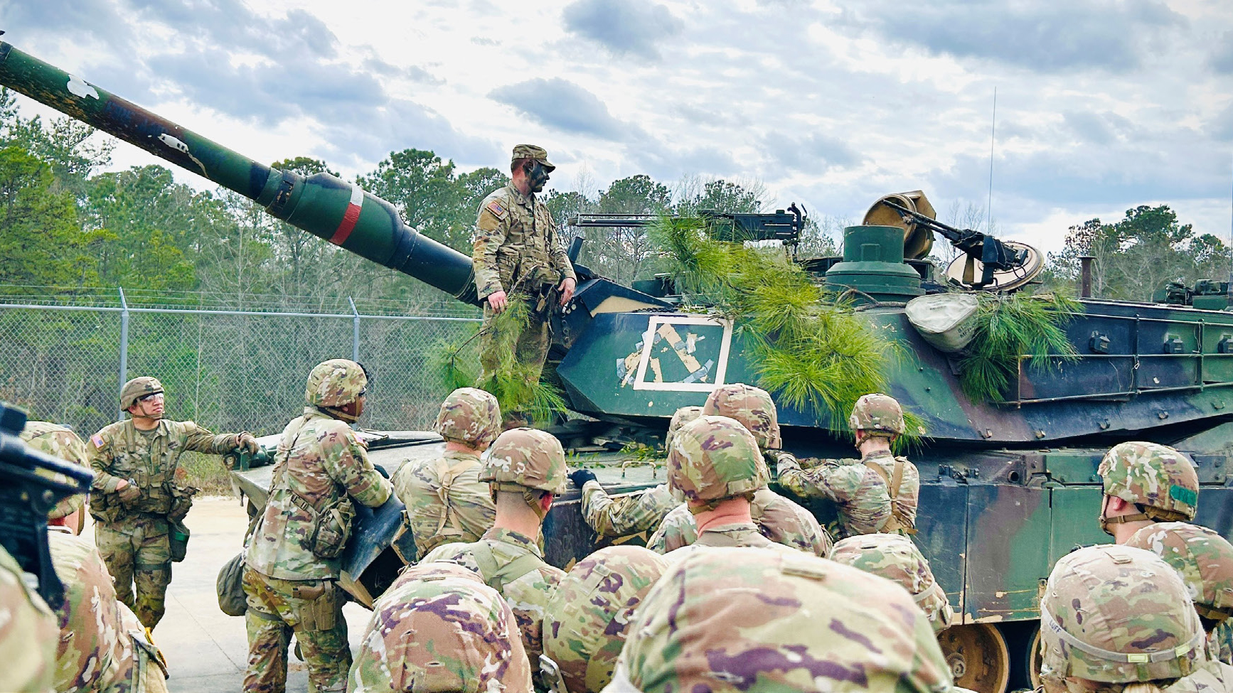 Group of U.S. Army soldiers in multicam uniforms gathered around a camouflaged self-propelled howitzer artillery piece during a training exercise, with trees and cloudy sky in the background