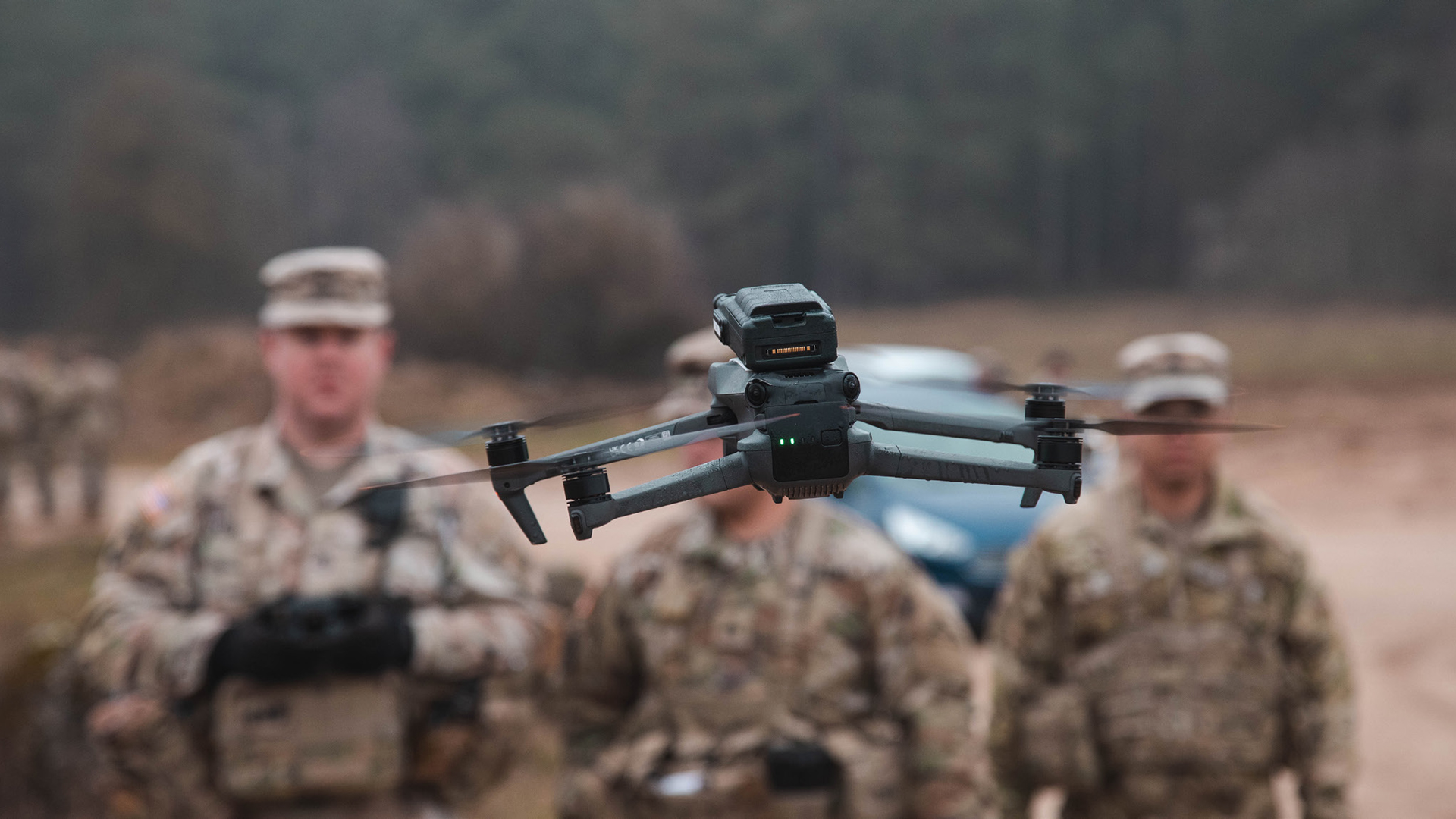 Gray military drone hovering in flight with green indicator lights, blurred U.S. Army soldiers in multicam uniforms visible in the background during field operations