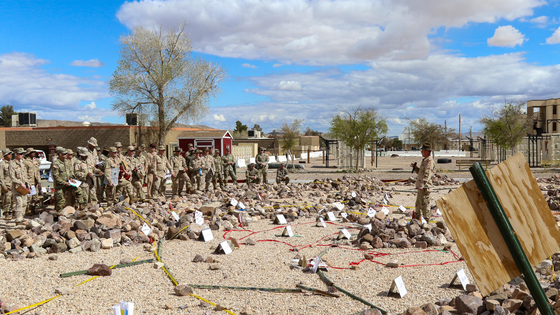 Military personnel in desert camouflage uniforms gathered around a training area filled with rocks and debris marked with numbered white cards and colored tape. The scene appears to be an outdoor explosive ordnance disposal or forensic training exercise under a cloudy sky with buildings visible in the background.