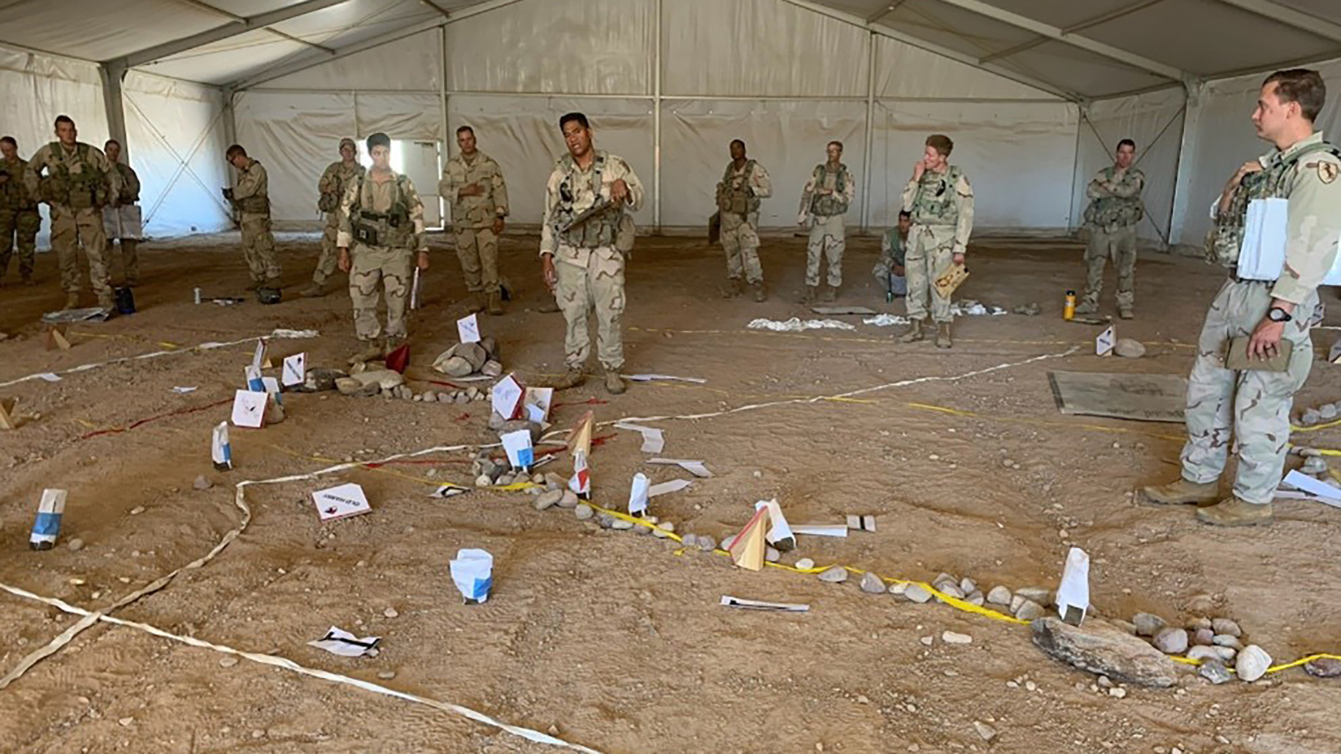 Military personnel in desert camouflage uniforms standing inside a large white tent observing a ground training exercise. The floor is covered with various objects, numbered markers, and colored tape laid out in a grid pattern, suggesting an evidence collection or bomb disposal training scenario.