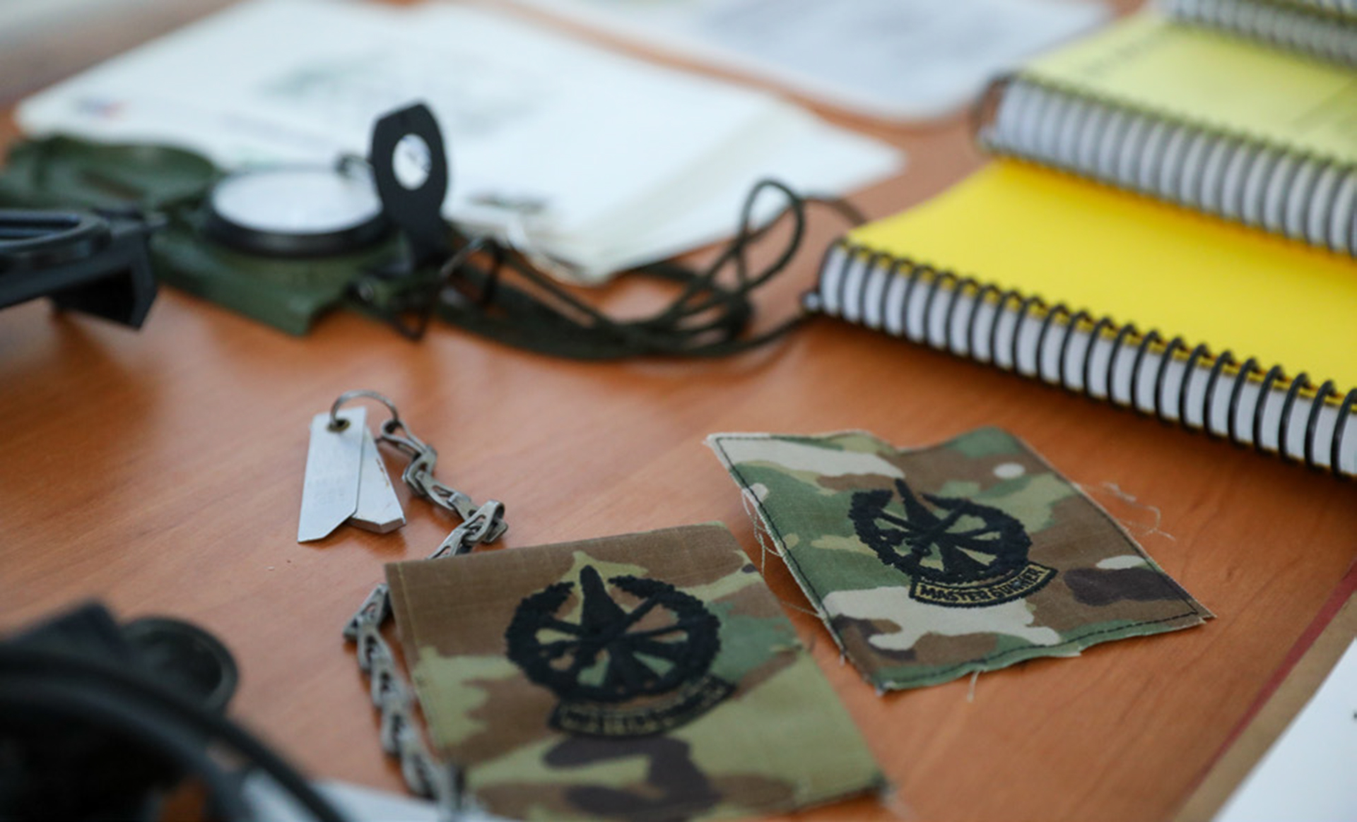Two camouflage military patches with artillery insignia lying on a wooden desk surface, alongside military dog tags on a chain. Yellow spiral notebooks and other military equipment are visible in the blurred background.