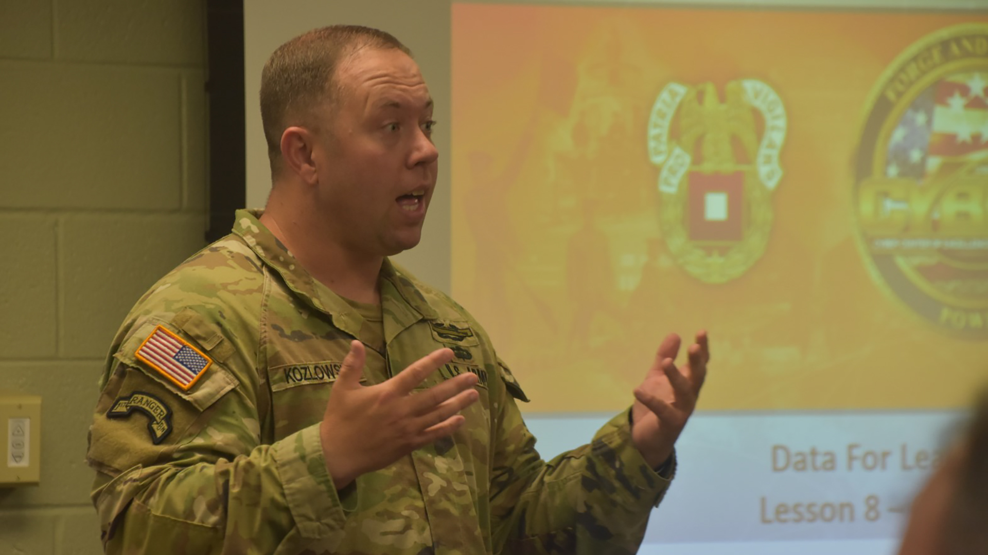 U.S. Army Chief Data Officer, Capt. Derrick Kozlowski, giving a presentation in a classroom setting. He is gesturing with his hands while speaking, wearing standard Army camouflage uniform with visible patches including an American flag. Behind him is a projection screen showing military insignia or emblems, with text partially visible at the bottom that reads 'Data for Le... Lesson 8'. The setting has institutional walls typical of a military or educational facility.