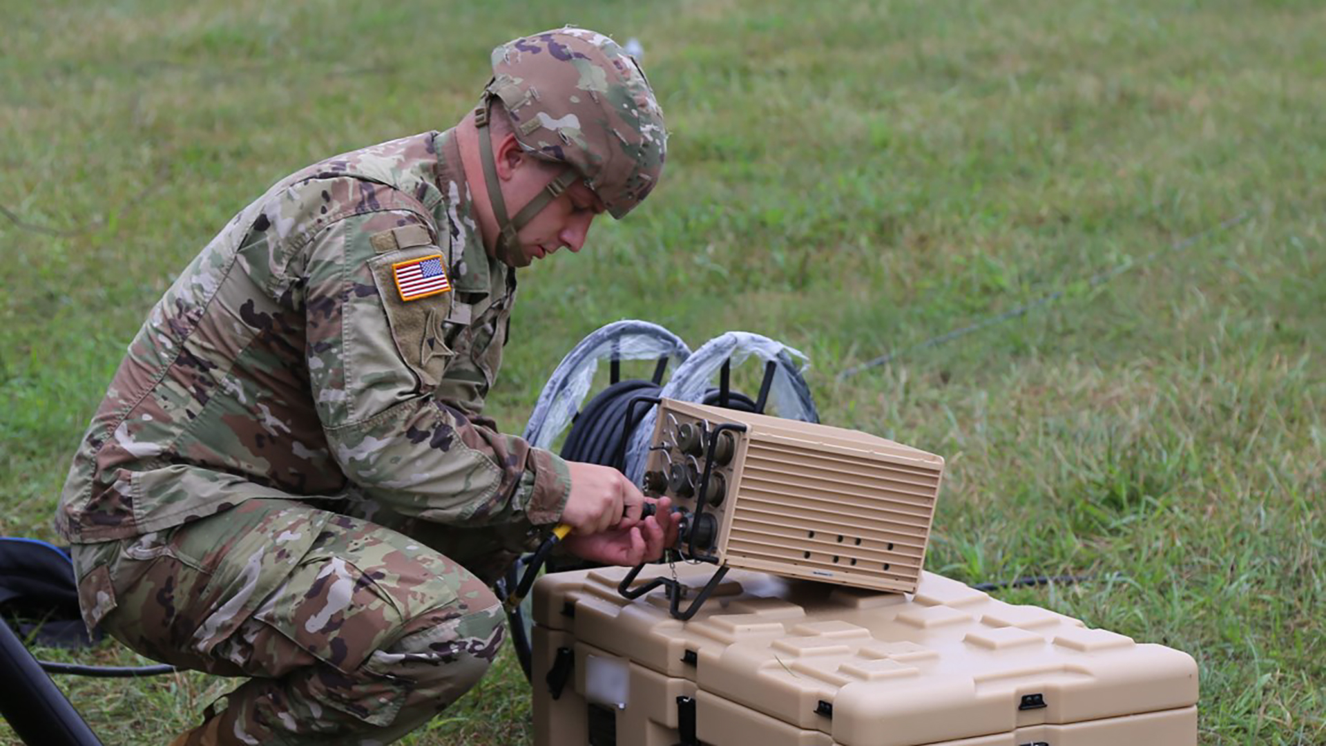 The image shows a soldier in camouflage combat uniform with an American flag patch visible on the sleeve, kneeling on grassy terrain. The soldier is operating or setting up what appears to be military communications equipment. There are two tan/beige tactical cases or containers on the ground - one larger storage case and what looks like a communications device or radio system with a control panel. This appears to be a field communications setup, likely part of a military training exercise or deployment where the soldier is establishing a tactical communications link.