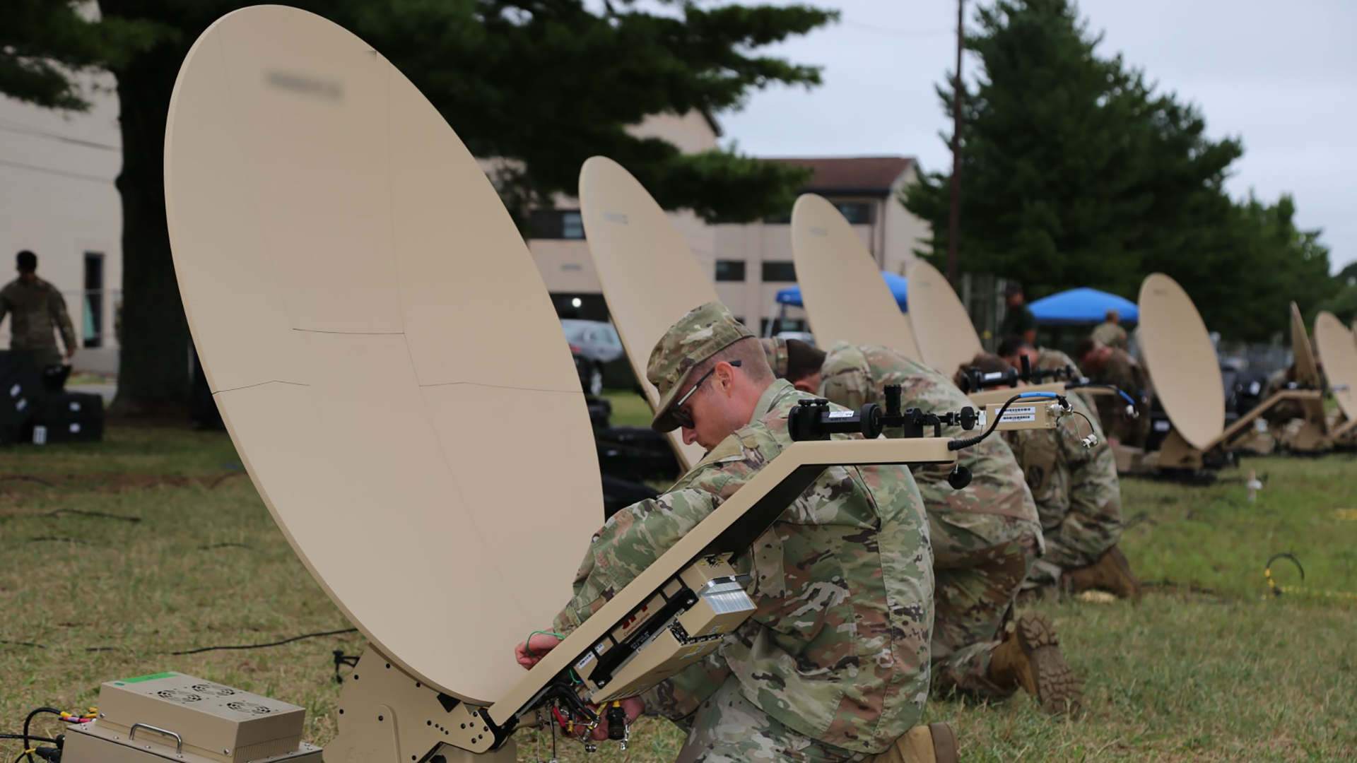 The image shows military personnel in camouflage uniforms setting up portable satellite communication systems in a field. Multiple soldiers are visible working with several deployed satellite dishes - these are portable VSAT (Very Small Aperture Terminal) systems with their distinctive white parabolic dishes positioned on the ground. The personnel appear to be establishing a tactical communications network, likely as part of a military exercise or deployment. In the background, additional satellite dishes can be seen along with what looks like temporary structures or tents. This represents field communications infrastructure being established for military operations, allowing for secure voice and data transmission capabilities in remote locations.