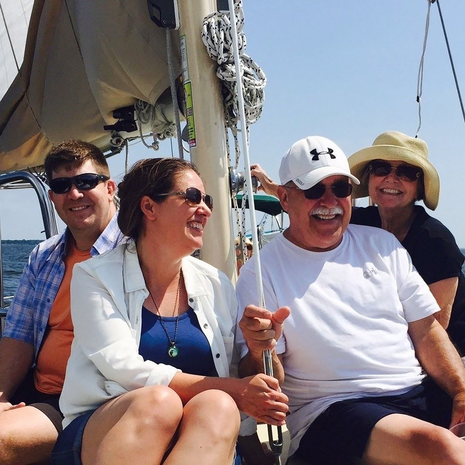 Four people enjoying a sunny day on a sailboat, all wearing sunglasses and summer clothing. They are seated together on the boat deck with the ocean visible in the background and sailing equipment like ropes and rigging visible around them.