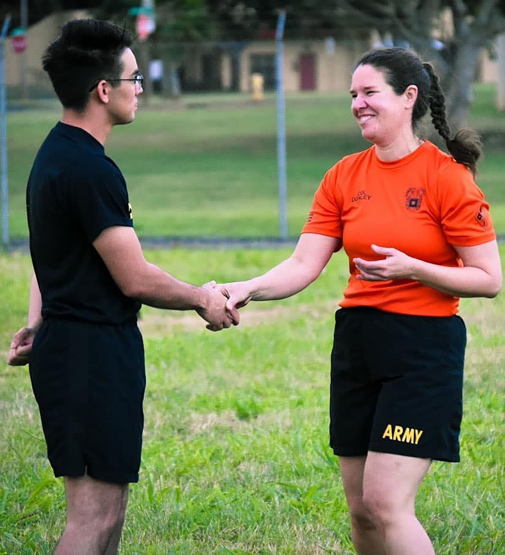 Two people in athletic wear shaking hands on a grassy field with goalposts visible in the background. One person wears a black shirt while the other wears an orange shirt and black shorts with ARMY printed on them.