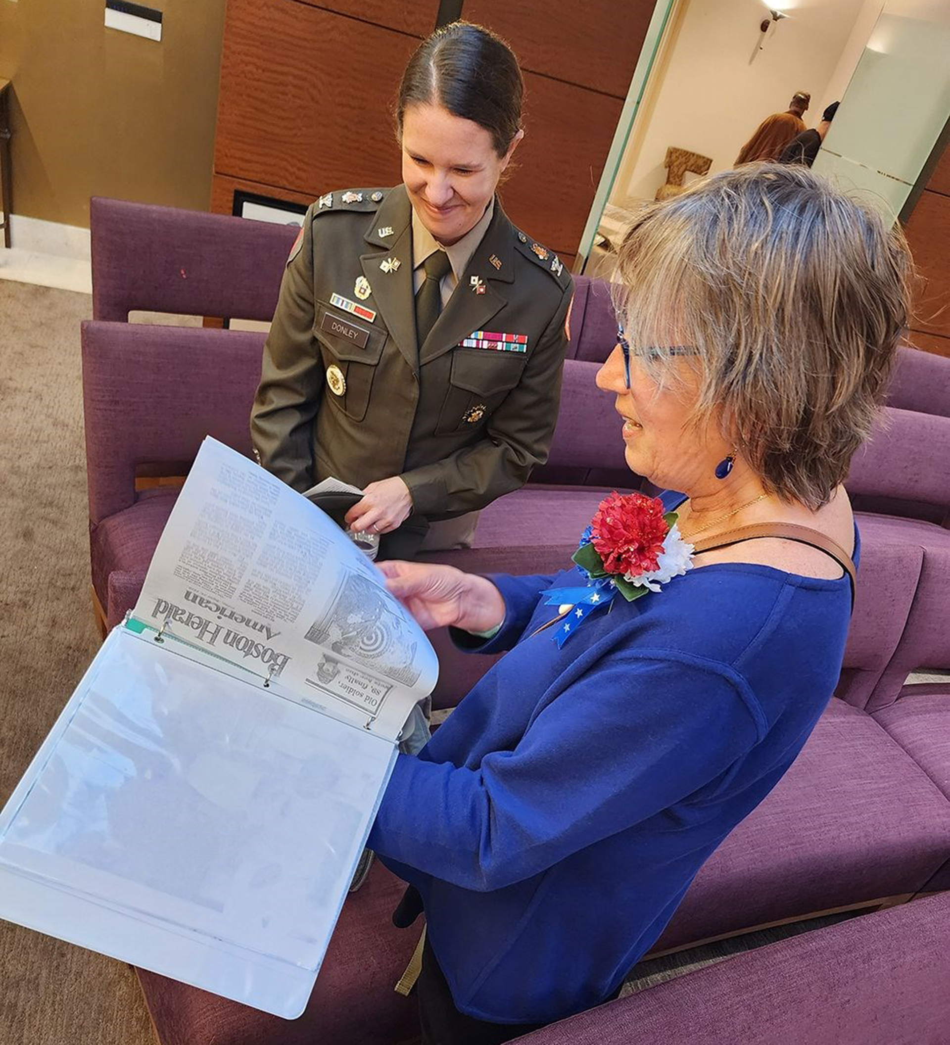 A female soldier in dress uniform sitting on a purple couch next to an older woman in a blue dress who is reading a newspaper. The woman in blue is wearing a red carnation corsage and the setting appears to be indoors