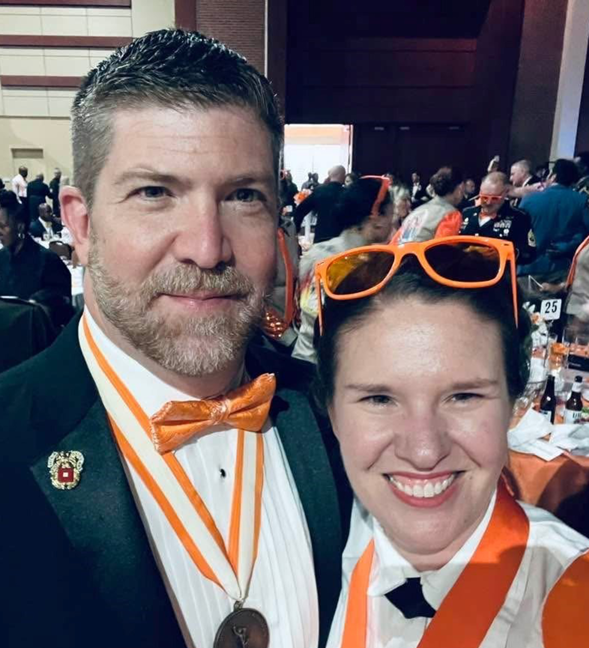 A man and woman at what appears to be a formal event or graduation ceremony, both wearing orange and white academic regalia. The man has an orange bow tie and medal, while the woman wears orange sunglasses on her head. They are smiling at the camera with a crowd visible in the background.