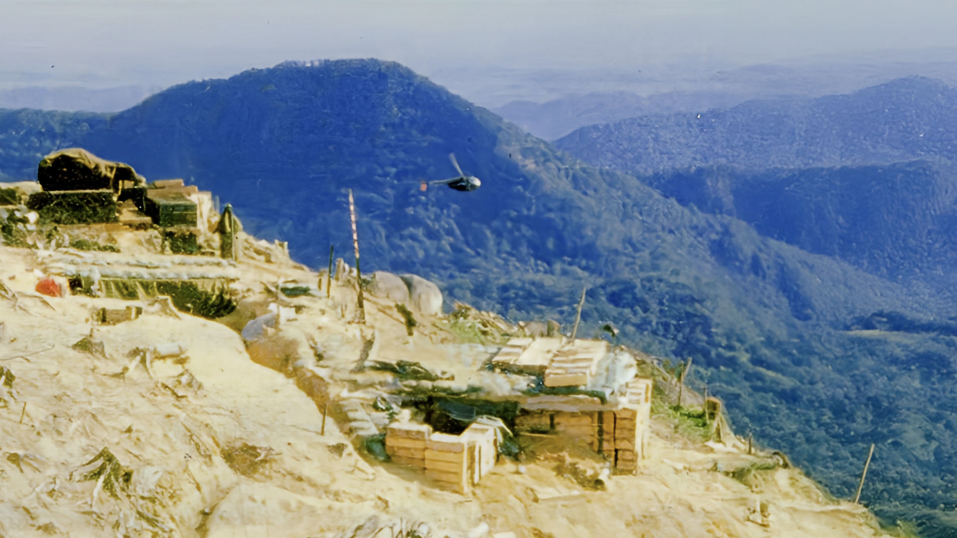 Aerial view of Fire Support Base Ripcord showing fortified positions, bunkers, and defensive structures on a hilltop with mountains in the background and a helicopter in flight
