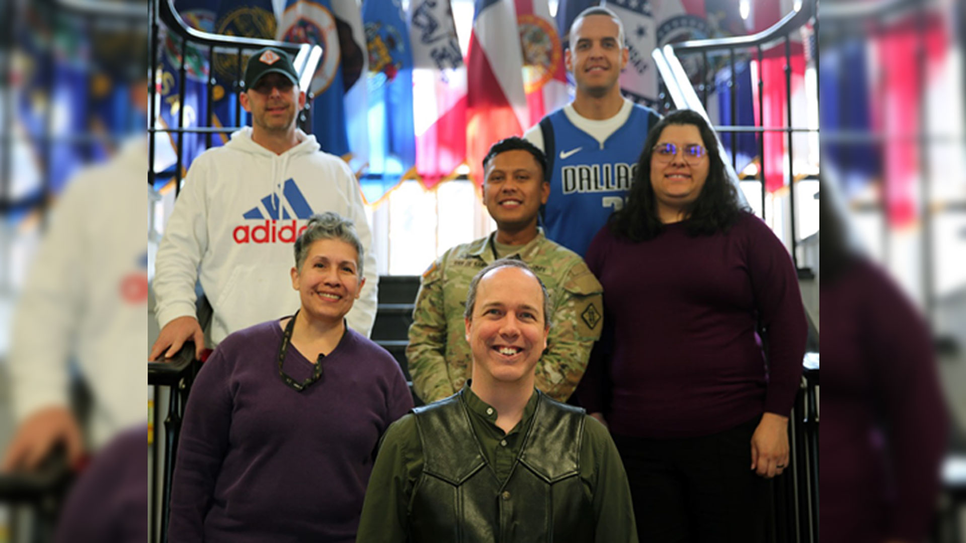 Group of six people posing for photo in front of colorful military unit flags display, including one person in Army camouflage uniform and five in civilian clothing including sports jerseys and casual wear.
