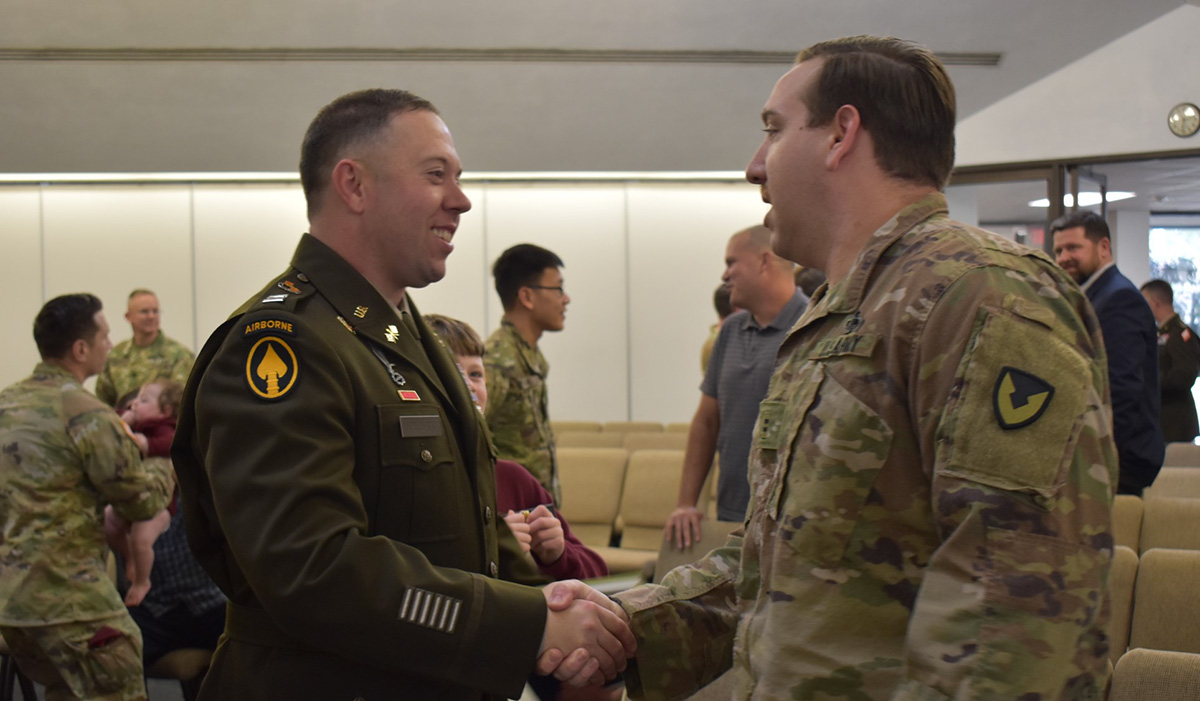 A newly commissioned U.S. Army Signal Corps officer in a green service uniform shakes hands with a soldier in an operational camouflage uniform during a ceremony. Other uniformed service members and civilians are visible in the background, engaging in conversations. The setting appears to be an indoor hall with beige seating and bright lighting.