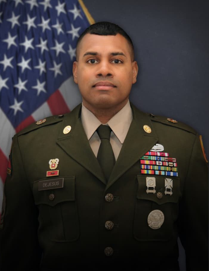 U.S. Army soldier in formal military dress, standing in front of the American flag. The soldier has a close-cropped haircut and wears a green service uniform jacket adorned with various badges, ribbons, and medals. The nameplate on the uniform reads 'DEJESUS.' The soldier's expression is composed and focused, and the backdrop is a dark gradient, highlighting the individual in the foreground.