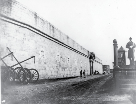 A historical black and white photograph of a fort’s seacost facing wall from 1875, showing soldiers standing near the entrance.