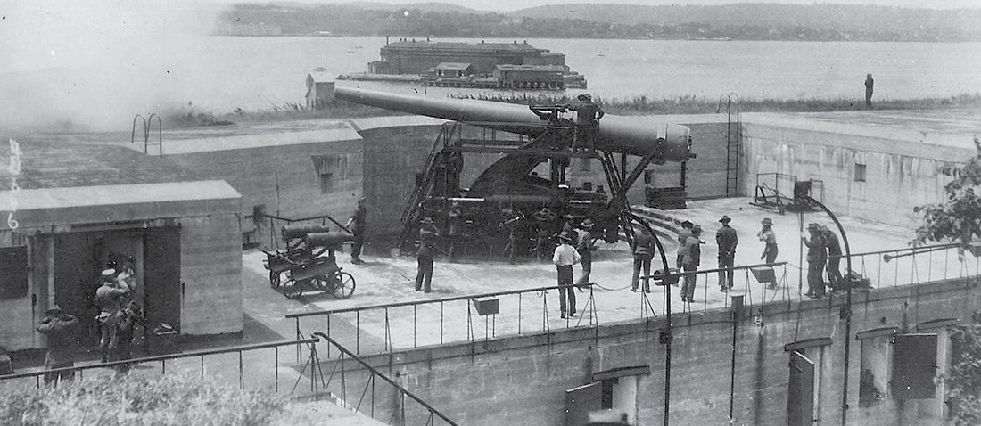 A historic military emplacement, with large guns and soldiers in uniform, with additional fortifications in the background.