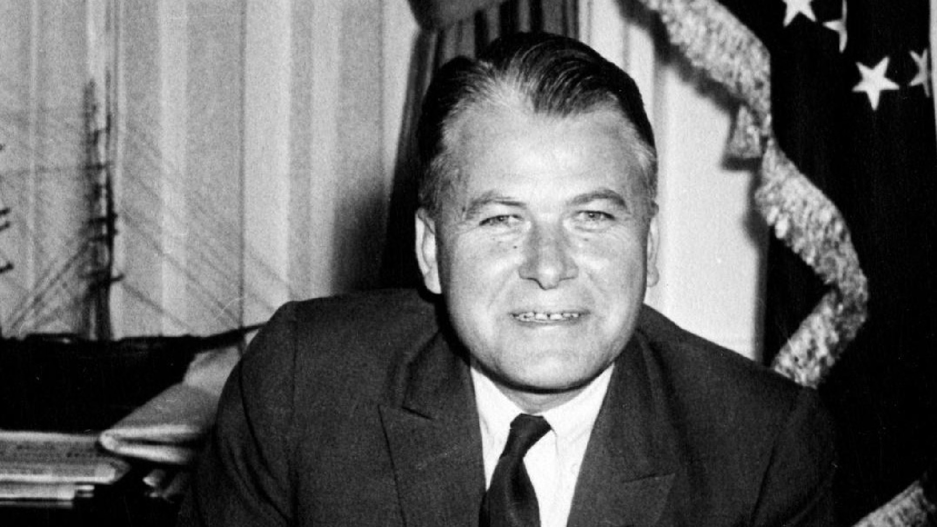 Black and white photograph of a middle-aged man in a dark business suit and tie, seated at a desk with his hands folded over papers. He has slicked-back hair and is smiling at the camera. Behind him are curtains and an American flag with decorative fringe, suggesting this is an official government office setting from what appears to be the mid-20th century based on the photographic style and office decor.