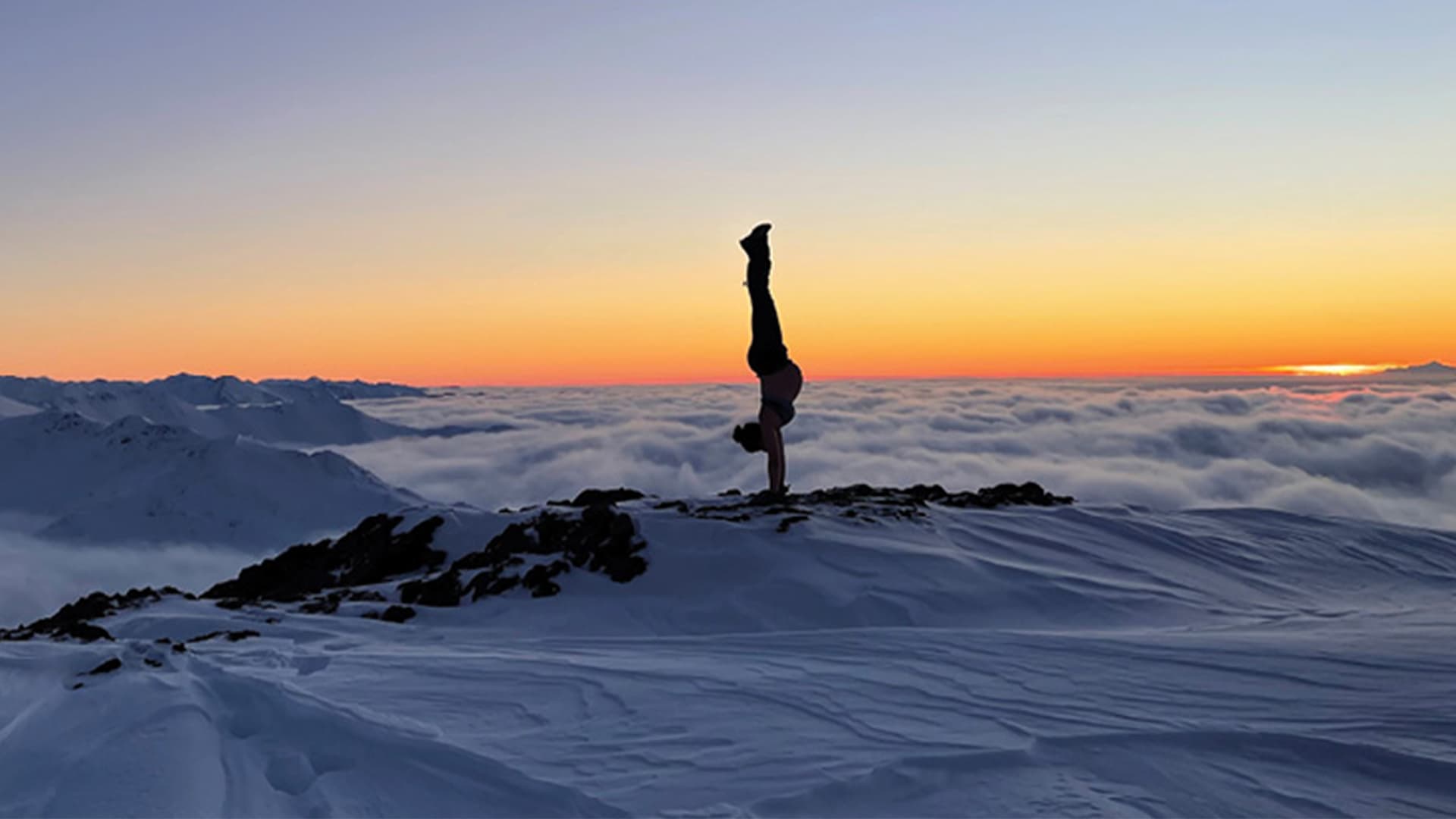 A person performing a handstand on a snowy mountain peak at sunset. The horizon is filled with soft, billowing clouds beneath the mountaintop, while the sky above transitions from deep blue to warm orange and pink hues as the sun sets. The scene is serene and awe-inspiring, with the person silhouetted against the vibrant sky, emphasizing both their balance and the vastness of the surrounding landscape.