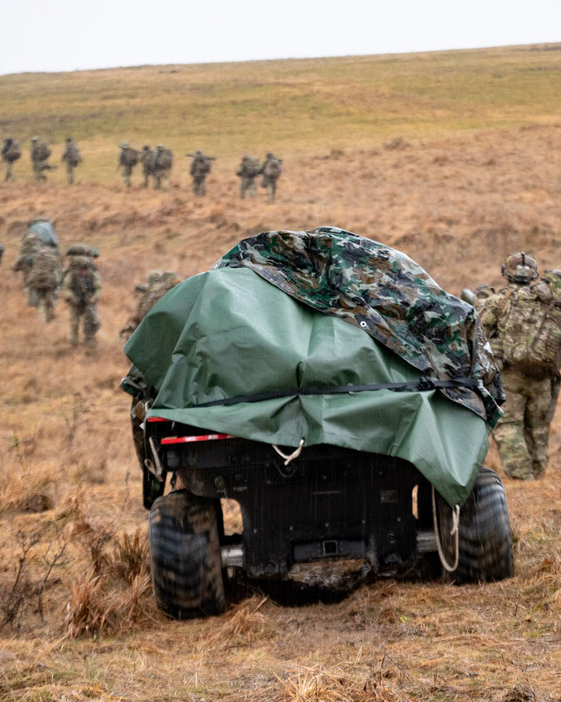 Military exercise scene with soldiers walking uphill across a grassy field while a covered utility vehicle with camouflage tarp is parked in the foreground.