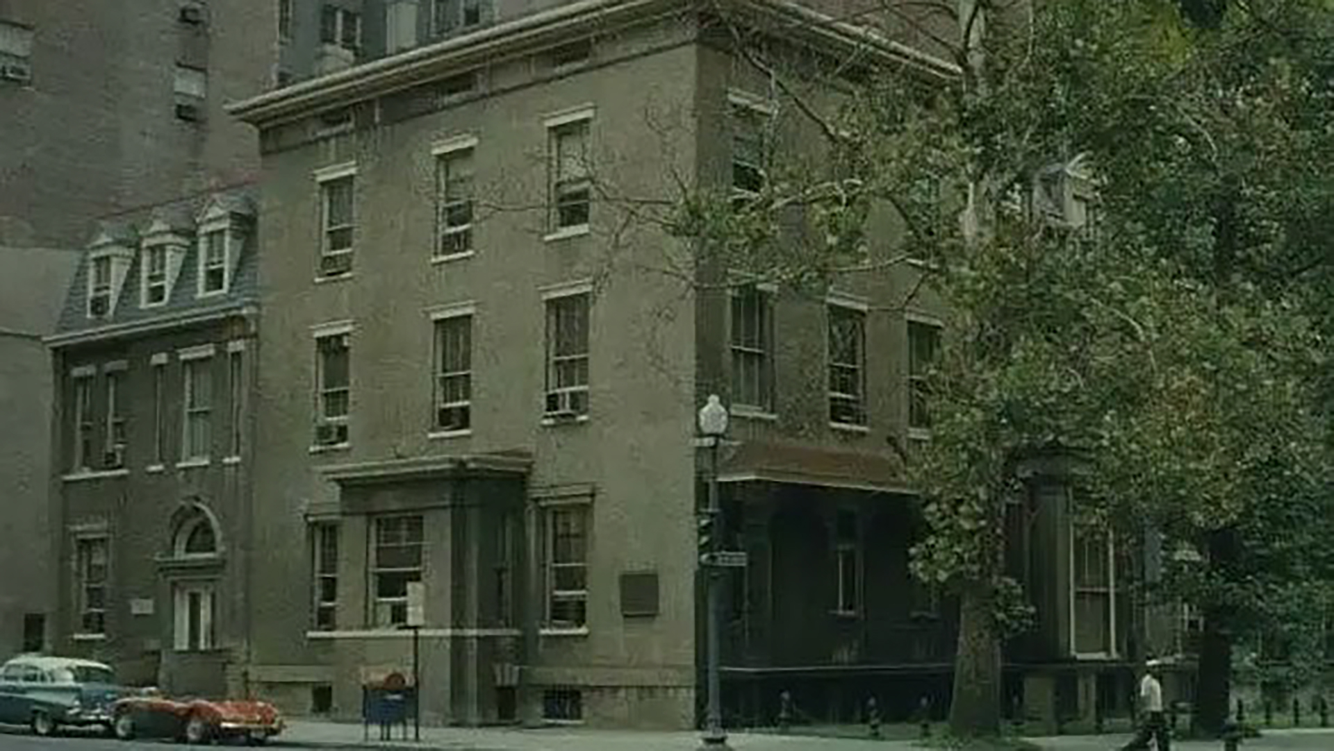Historic three-story brick townhouse on a city street corner with trees, parked cars, and tall apartment buildings in the background.