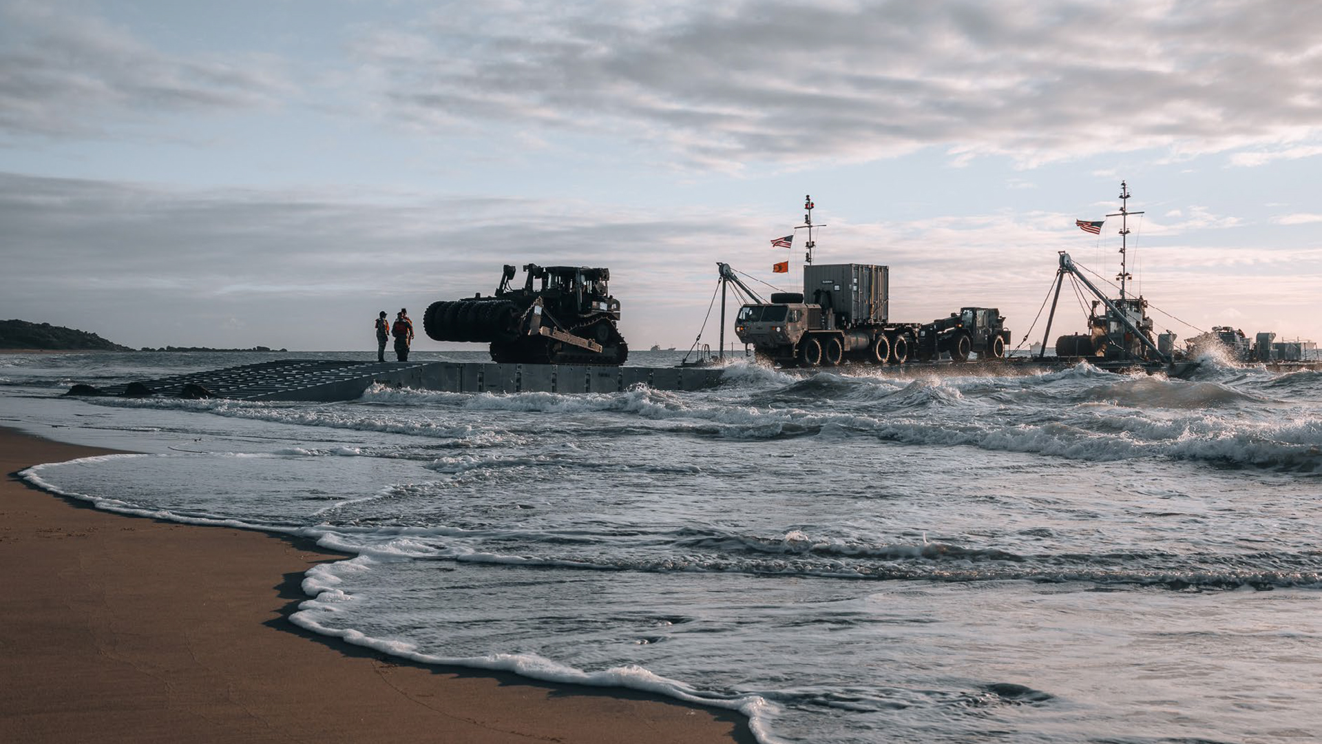 A coastal scene showing military vehicles and equipment on a floating platform near the shoreline. Two service members stand on the platform, overseeing operations. A bulldozer and other heavy machinery are visible, with waves crashing against the platform. The background features a cloudy sky and calm waters, emphasizing the logistical operation in a maritime environment.