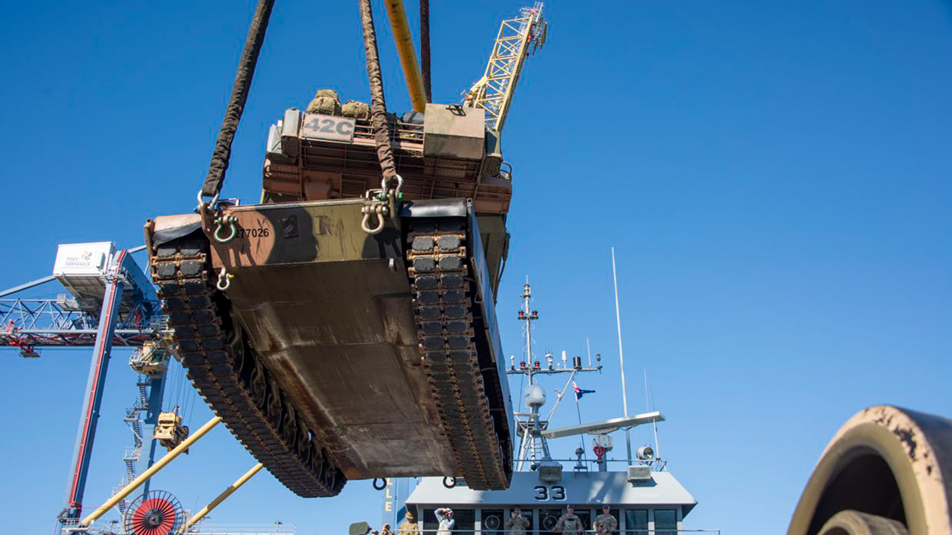 A military tracked vehicle being lifted by crane cables above a naval vessel. The vehicle appears to be an armored personnel carrier or similar military equipment marked with identification numbers. Naval personnel are visible on the ship's deck below, and the operation is taking place under clear blue skies.