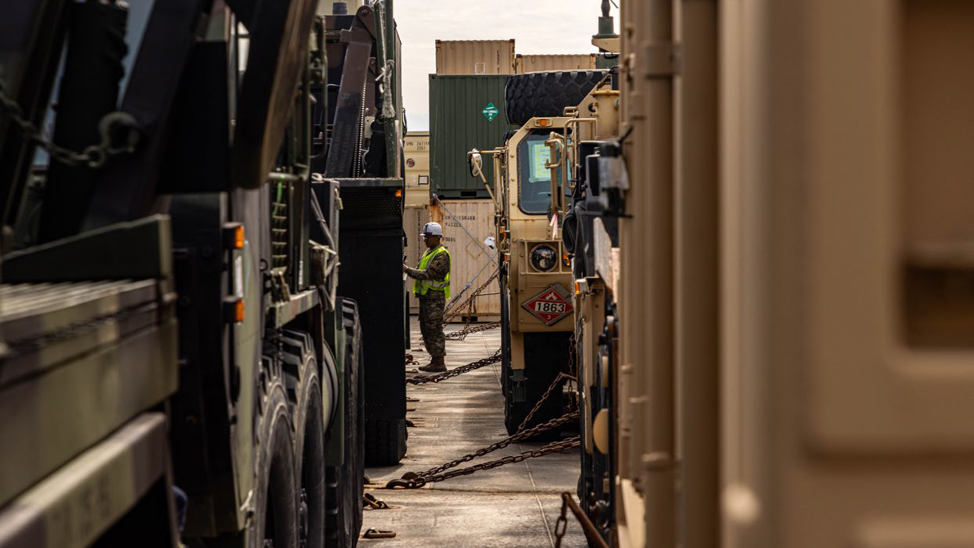View down a narrow corridor between military vehicles showing heavy trucks and equipment lined up. A soldier in high-visibility safety gear stands among the vehicles. The scene appears to be at a military base or staging area with shipping containers visible in the background.